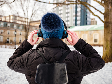 Female university student with headphones, listening to favourite tunes. Exterior of city park with historic buildings in the background.