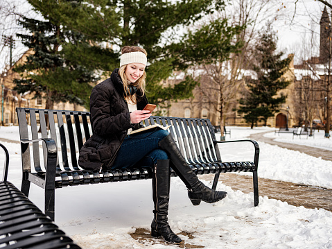 Female university student reading her notes while sitting on the park bench. Exterior of city public park in winter  with historic buildings in the background.