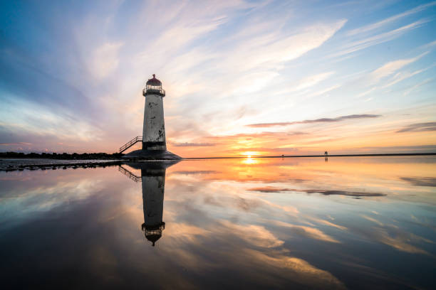 水の見事な日没の日の出反射のプールに立っている灯台は、北ウェールズの海岸砂浜を構築するために水と海のステップに反映され、まだ水オレンジ色の輝く黄金の時間の青い時間 - 静水 ストックフォトと画像