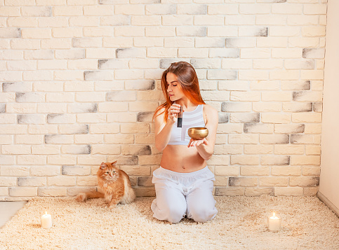 A young sports girl meditates using a Tibetan singing bowl. A beautiful woman sits at home on a carpet next to a cat and plays on a singing bowl. Meditation and healthy lifestyle concept. Copy space.