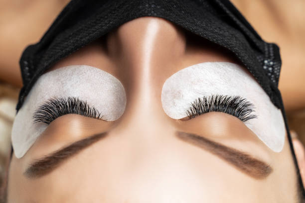 istock Eyelash Extension Procedure. Beautiful Woman with Extreme Long False Eyelashes. Makeup, Cosmetics. Beauty, Skincare. Woman in medical mask 1300016561