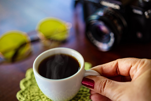 Goiania, Goias, Brazil – February 02, 2021: Female hand holding a cup of coffee, with a camera and yellow lens glasses on a wooden table.