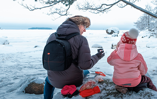 Family of Caucasians on a walking tour, picnic on the shore of the bay in winter. Dad and daughter drink hot tea from a thermos and enjoy nature.