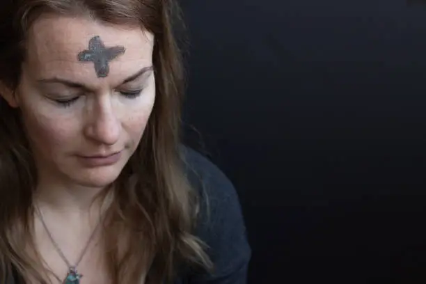 Caucasian woman with ash cross on forehead for Ash Wednesday looking down with eyes closed with black background and copy space