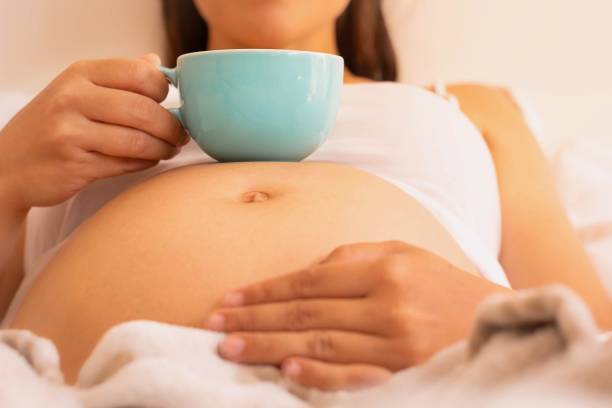 Pregnant woman drinking a cup of coffee and relaxing at home. A close up of a pregnant baby belly, woman holding a mug with coffee or tea, and laying in bed. hot filipina women stock pictures, royalty-free photos & images