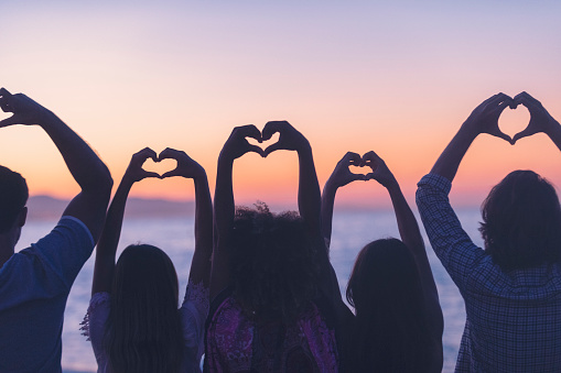 Group of young people making love heart signs at sunset or sunrise. They are facing away from the camera and are in silhouette.