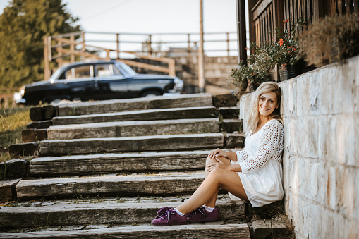 Beautiful young woman with long blond hair sitting and relaxing on wooden stairs in front of a house on sunny summer day