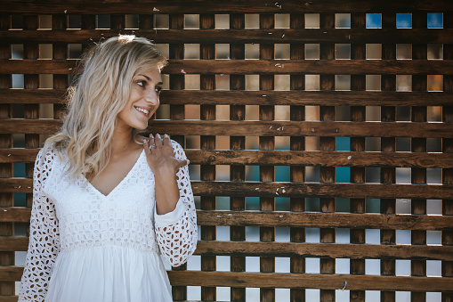 Beautiful young woman with long blond hair standing and leaning on brown wooden fence outdoors on sunny summer day