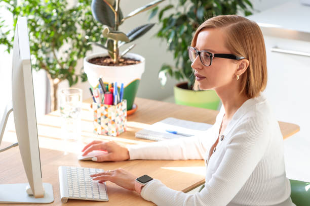 istock Pensive freelancer woman in glasses working on computer from home office. Cozy workplace surrounded by plants. Online business with distance job. 1300035607