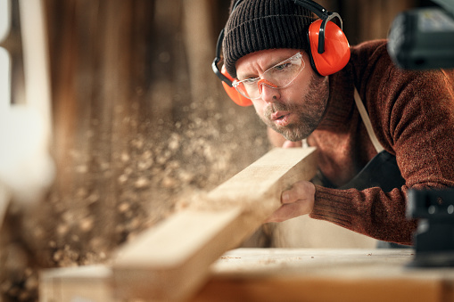 Adult male woodworker in protective goggles and headphones blowing sawdust from wooden detail while working in carpentry workshop