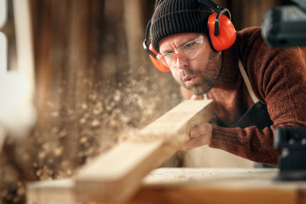 Carpenter blowing sawdust from wooden plank Adult male woodworker in protective goggles and headphones blowing sawdust from wooden detail while working in carpentry workshop carpentry photos stock pictures, royalty-free photos & images