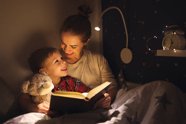 Cheerful mother and son cuddling and reading book Happy family woman and boy cuddling and smiling while reading book on bed near lamp at night at home bedtime stock pictures, royalty-free photos & images