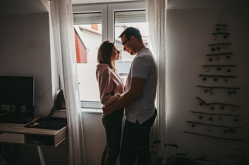 Young romantic Caucasian couple standing and hugging by the window during staying at home order, while their cat sitting on window sill