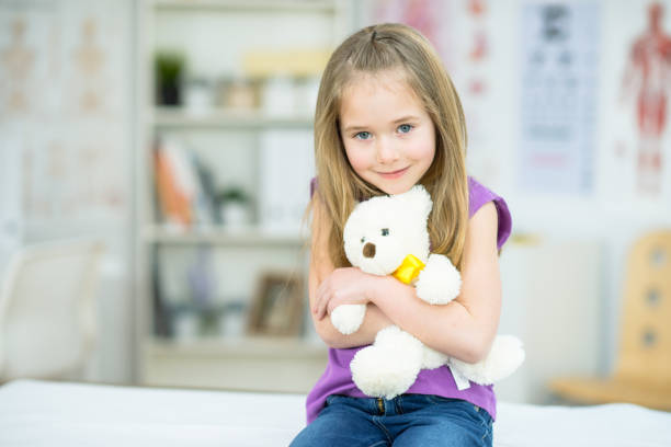 A super cute Caucasian girl around 4 years old is sitting on an examination table at a doctor's office. She is holding her teddy bear close to her chest and smiling at the camera.