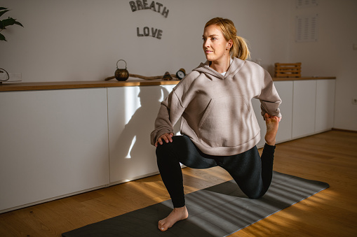 Full length view with sunlight and shadows of mid adult Caucasian woman in sweatshirt and yoga pants on exercise mat in Anjaneyasana pose.