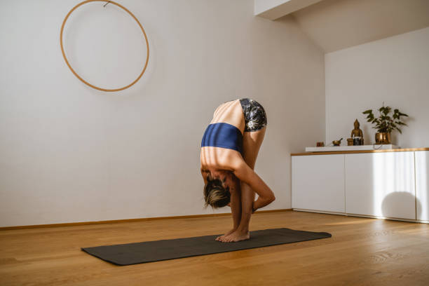 Full length side view of Caucasian woman wearing tube top and shorts in Uttanasana pose on exercise mat in yoga studio.