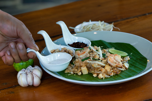 A Thai chef has prepared a delicious traditional Thai street food cuisine called ‘Pad Thai’ served with prawns.  He lays side dishes of Chili, sugar, lime and peanut on the side.