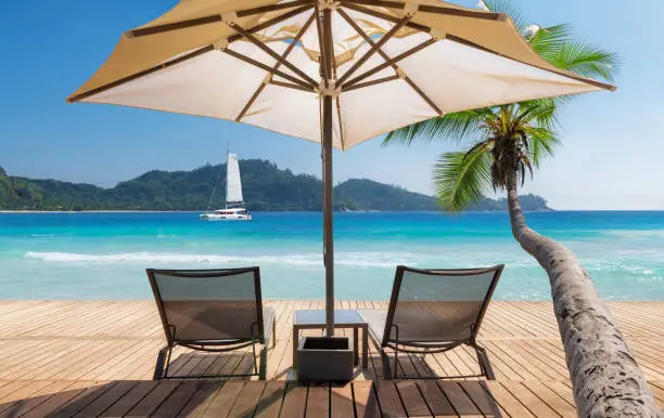 Sunny beach with umbrella and deckchairs on wooden floor, coconut palm trees and a sailing boat in the turquoise sea on Paradise island in tropical ocean. Summer vacation and tropical beach concept.