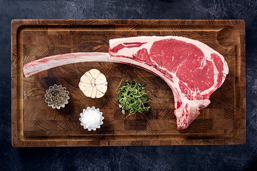 Overhead view of a huge, raw tomahawk steak being prior to being cooked. With; salt, pepper, a whole garlic bulb halved and some fresh thyme. Colour, horizontal with some copy space. The Tomahawk Steak got it’s name from their shape resembling a tomahawk axe. They have become popular in restaurants and with home barbecue chefs to see how much one person can eat!