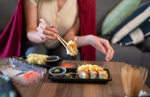 Woman having takeaway or delivery sushi rolls meal from a plastic package with sauces sitting on the sofa covered with a blanket for a cozy feeling at home