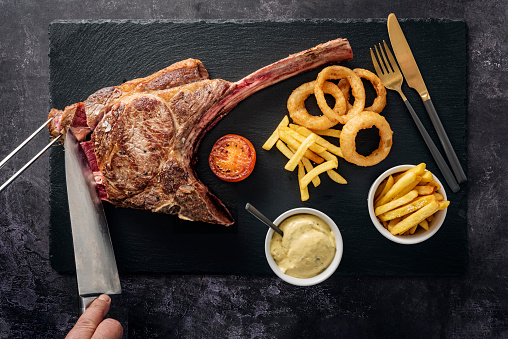 Overhead view of a huge, cooked tomahawk steak being served with; french fries, tomato slice, deep fried onion rings and béarnaise sauce as it is sliced at the table. Colour, horizontal with some copy space. The Tomahawk Steak got it’s name from their shape resembling a tomahawk axe. They have become popular in restaurants and with home barbecue chefs to see how much one person can eat!