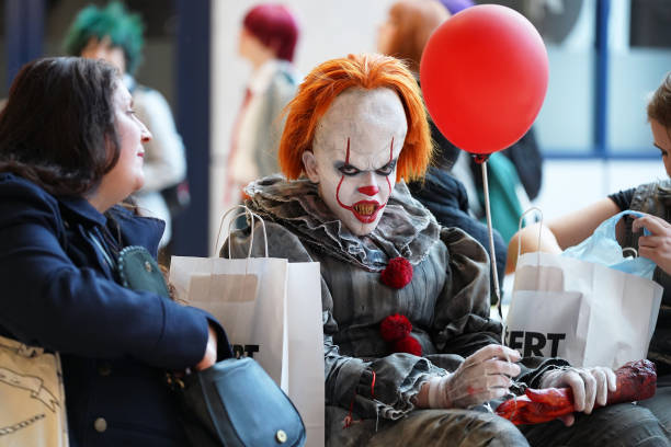 Birmingham NEC, England, UK - 24th March 2019\n\nScary horror clown Pennywise with orange red hair, large forehead and sinister makeup holding a floating balloon. Sat with general public giving a frightening evil stare to the camera.