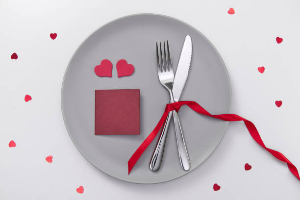 Valentine's day festive table setting card for text, flat lay with red heart shape on gray plate, fork, knife and red ribbons on white table. Love dating concept. Place your text and copy space