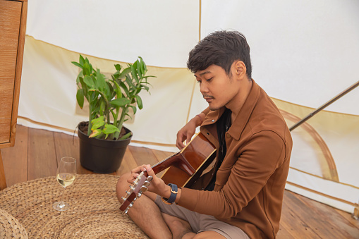 Candid shot of an Indonesian man playing guitar in a luxurious white tent while glamping with a glass of white wine in front of him.