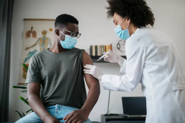 Young African American male patient sitting in a medical clinic and is being given the Covid 19 vaccine in his shoulder by a female African American doctor, both wearing protective face masks
