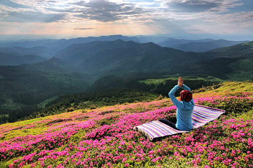 Yoga girl is sitting on the striped mat in meditation on the lawn with pink flowers at the high mountains and sky with rays and clouds. Beautiful spring morning.