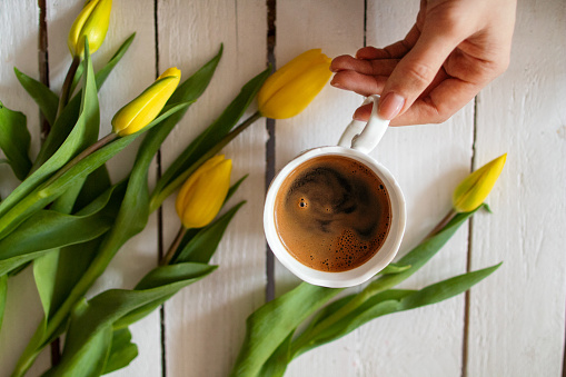 Beautiful yellow tulips with bright green leaves laying on the white wooden table. Fresh hot coffee with a bubbly foam is making a statement in this beautiful morning ritual scene. Human hand is holding a coffee mug, ready for the first sip.