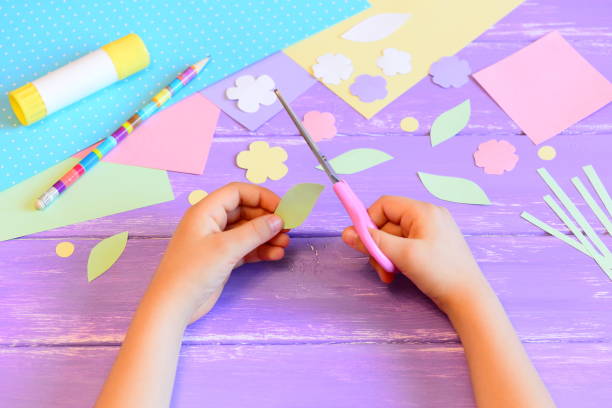 istock Small child does a greeting card for mom. Step. Kid holds scissors in hand and cuts a leaf from paper. Colored paper, paper templates, glue stick, pencil on a table. Mother's day or March 8 crafts diy 1300120959
