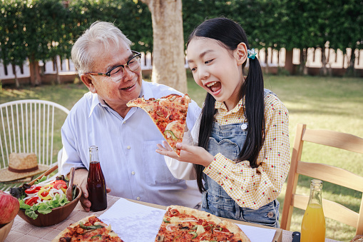 Asian retirement grandfather and pretty granddaughter enjoying to eating pizza together in home garden. Happy senior life after retirement with family concept.