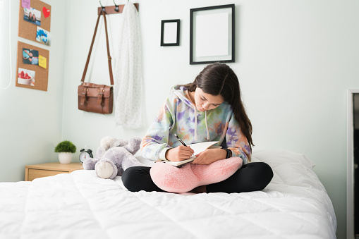 Thoughtful young teen girl in her bedrrom writing about her day on a diary while sitting in bed. Beautiful teen girl pouring her dreams and thoughts in a notebook