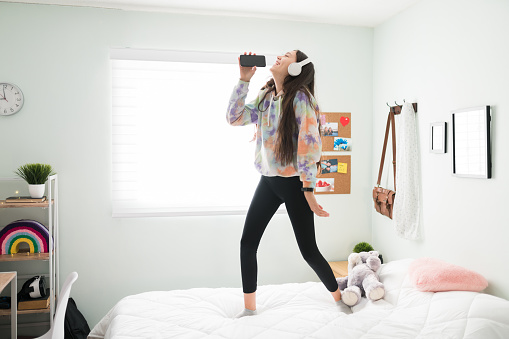 Playful and happy teenage girl having fun alone in her bedroom. Beautiful adolescent girl with headphones listening to music and singing along while standing on her bed