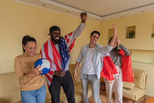 Four Multi-Ethnic Young Friends are Spending a Nice Time Together in Their Comfortable Apartment, Watching a Soccer Game, and Cheering Using an American and Canadian Flag.