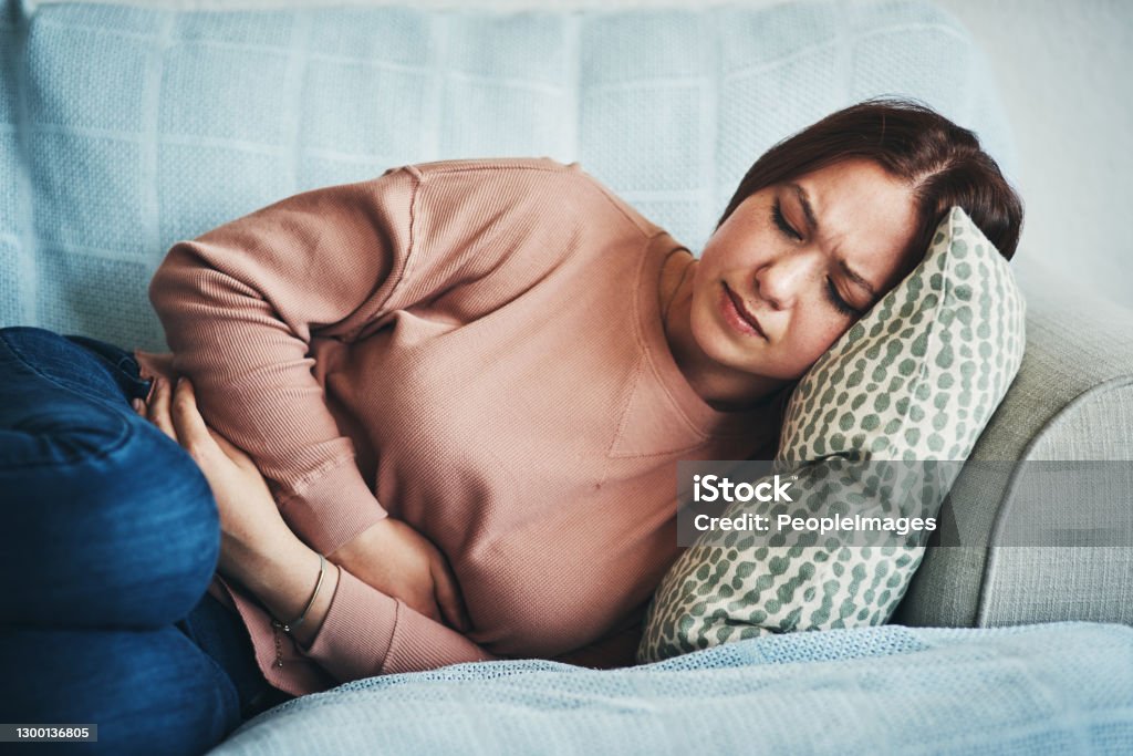 My gut doesn't feel so good Shot of a young woman experiencing stomach pain while lying on the sofa at home Stomachache Stock Photo