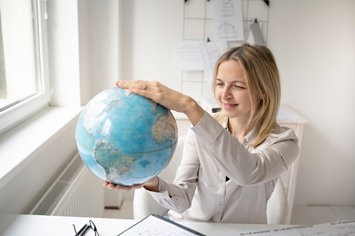 young, beautiful female architect is sitting in her office and is holding a globe and is dreaming of travelling around the world