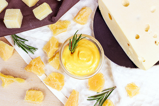 Top view of fresh homemade creamy cheese sauce in a glass bowl with rosemary and parmesan pieces on light background.