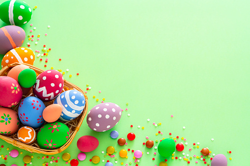 Overhead view of a group of hand painted colorful Easter eggs arranged in a wicker basket at the left and bottom of a green background making a frame and leaving useful copy space for text and/or logo. Sugar sprinkles and candies complete the composition. High resolution 42Mp studio digital capture taken with Sony A7rII and Sony FE 90mm f2.8 macro G OSS lens
