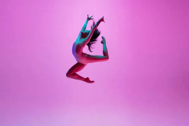High above. Young and graceful ballet dancer on pink studio background in neon light. Art, motion, action, flexibility, inspiration concept. Flexible caucasian ballet dancer, moves in glow.