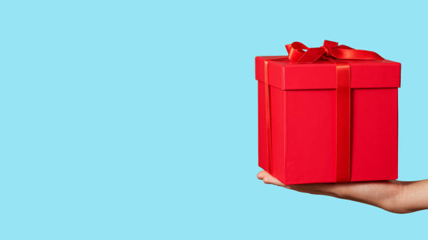 istock Hand holding red gift box with ribbon bow isolated on light blue background. Holiday concept. Copy space. 1300157797