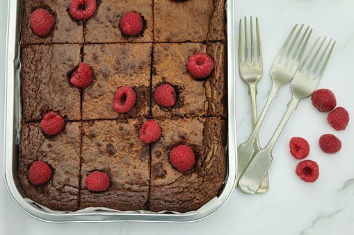 Stock photo showing elevated view of disposable tinfoil baking tray containing homemade chocolate and raspberry brownies cut into square slices, besides red raspberries and metal forks on a grey and white marble effect work surface.