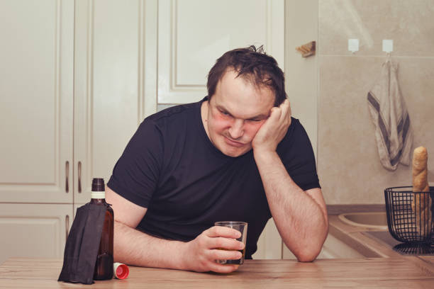 l’homme fâché boit l’alcool se reposant avec une bouteille de whiskey à la table de cuisine. problèmes d’alcoolisme pendant l’épidémie, l’isolement et le verrouillage - eastern european caucasian one person alcoholism photos et images de collection