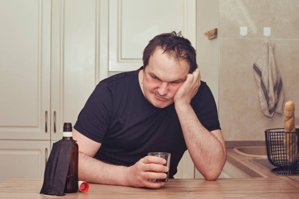 怒っている男は台所のテーブルでウイスキーのボトルと一緒に座ってアルコールを飲みます。流行、孤立、ロックダウン中のアルコール依存症の問題 - eastern european caucasian one person alcoholism ストックフォトと画像