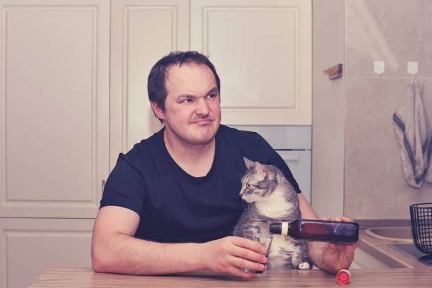男は思慮深く猫と台所に座ってアルコールを飲み、問題を振り返ります。絶望、悲しみ、憂鬱なアルコール依存症 - eastern european caucasian one person alcoholism ストックフォトと画像