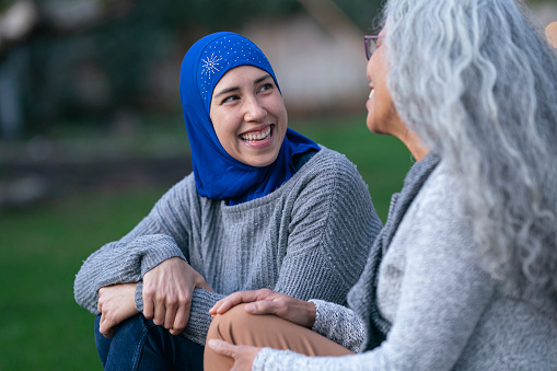 A beautiful muslim adult female in her 30's is wearing a hijab and spending time with her non-muslim mother outdoors. The mother has long, curly grey hair and they are both happy.