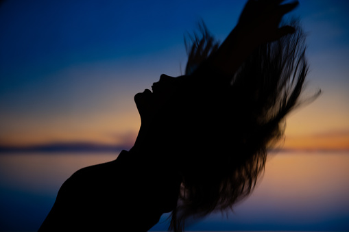 Inspirational and carefree moment of a woman throwing hair in the air in blue and orange sunset.