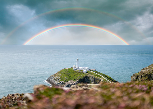 The beautiful North Wales South Stack lighthouse on a green island sat in a perfect blue sea with a double rainbow in the sky overhead.