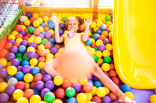 A funny girl sits in a playground with soft and bright equipment and throws colorful balls towards the camera while enjoying the warm summer sun